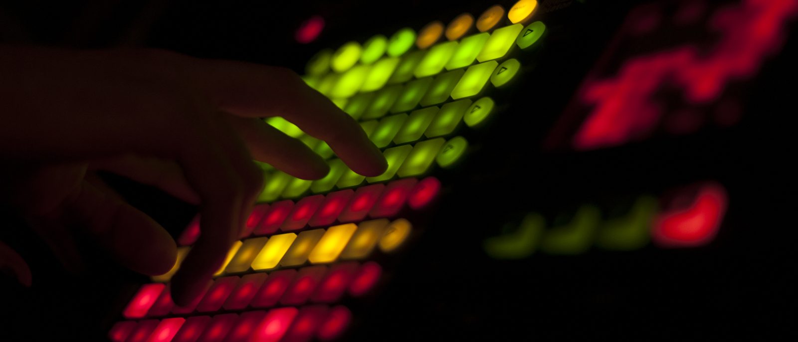 A hand playing a Novation Launchpad, pads lit red, yellow, and green in a dark room